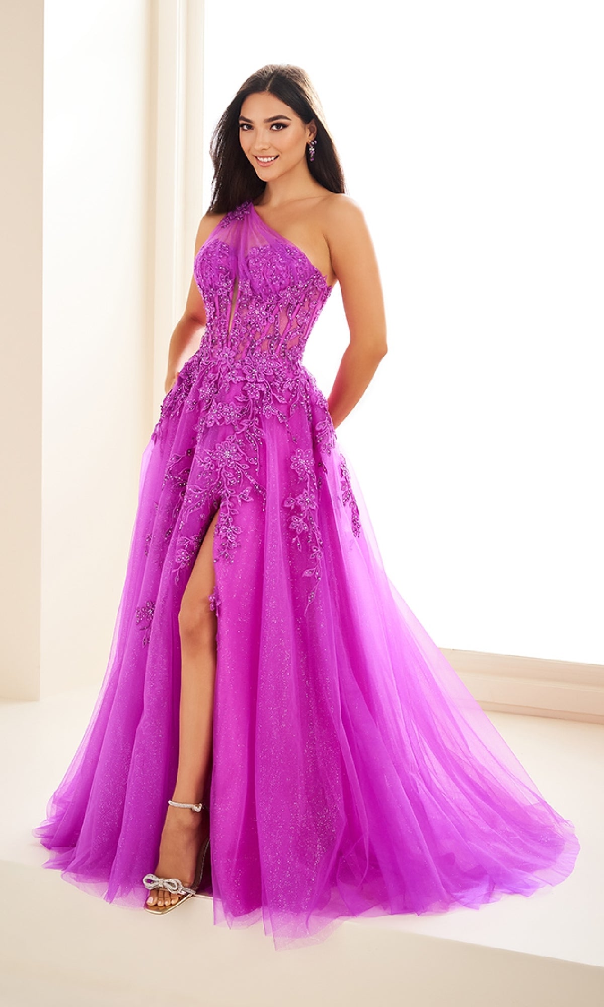 A woman wearing the Ellie Wilde EW36207 long prom dress—vibrant purple, one-shoulder, with floral embellishments, a corset bodice, and thigh-high slit—stands indoors by a bright window in high-heeled sandals.